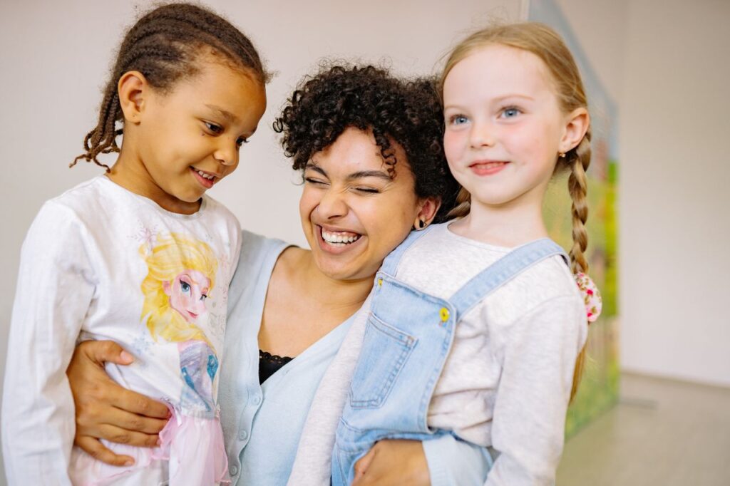 A joyful teacher hugging two smiling girls of different ethnicities indoors.
